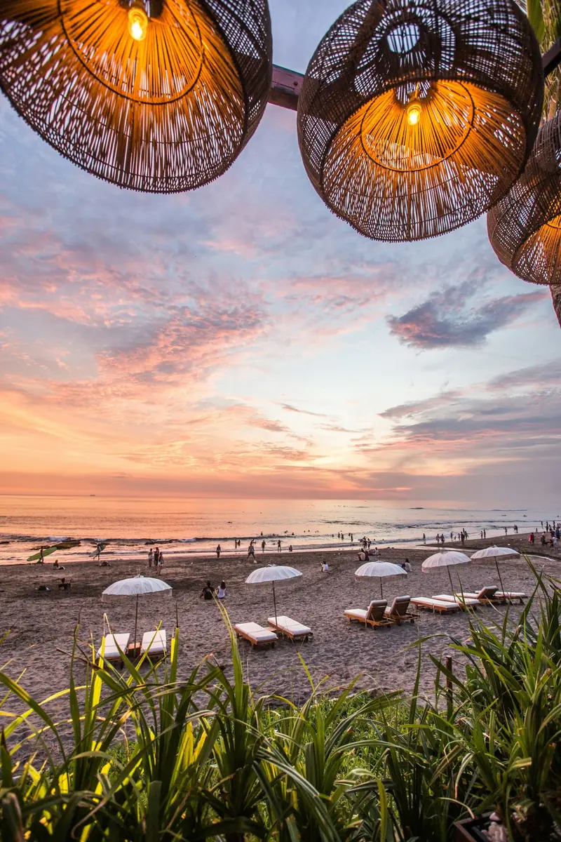 Sunset over a beach, with woven lamps and people along the shoreline.
